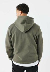 Haut à capuche vert olive avec cordon de serrage, fabriqué dans un tissu doux. Présente une coupe décontractée et un look superposé sur une chemise blanche.