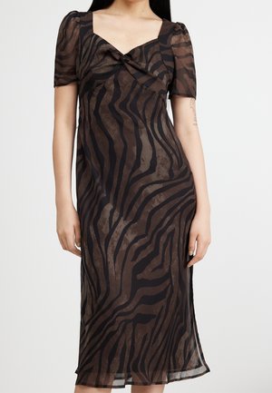 Kobieta ubrana w brązową przezroczystą sukienkę midi z czarnymi nieregularnymi paskami zebry, z bufkami na krótkich rękawach i zdobieniem w postaci skręconego węzła przy dekolcie.