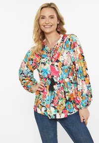 Blumenbluse mit langen Ärmeln, mehrfarbigem Blumenmuster, tailliertem Bund und Quasten-Detail am Ausschnitt, kombiniert mit blauen Jeans.
