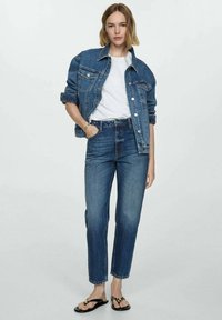 Jeansjacke in Mittelblau, weißes T-Shirt, hochgeschnittene blaue Jeans und schwarze Flip-Flops mit goldenen Akzenten. Model steht vor einem neutralen Hintergrund.