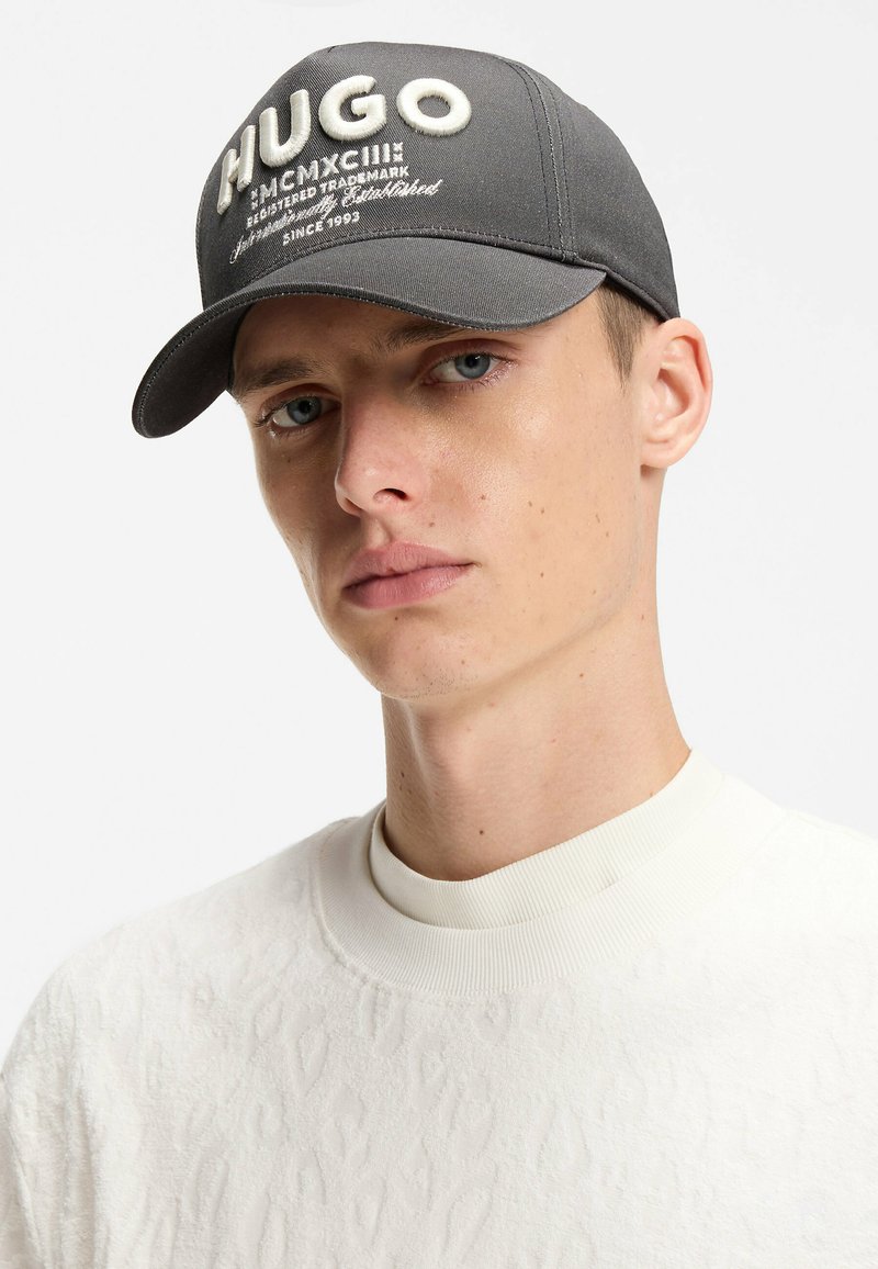 Jeune homme à la peau claire et aux yeux bleus portant une casquette HUGO gris foncé et un pull blanc texturé à col rond, sur fond blanc.