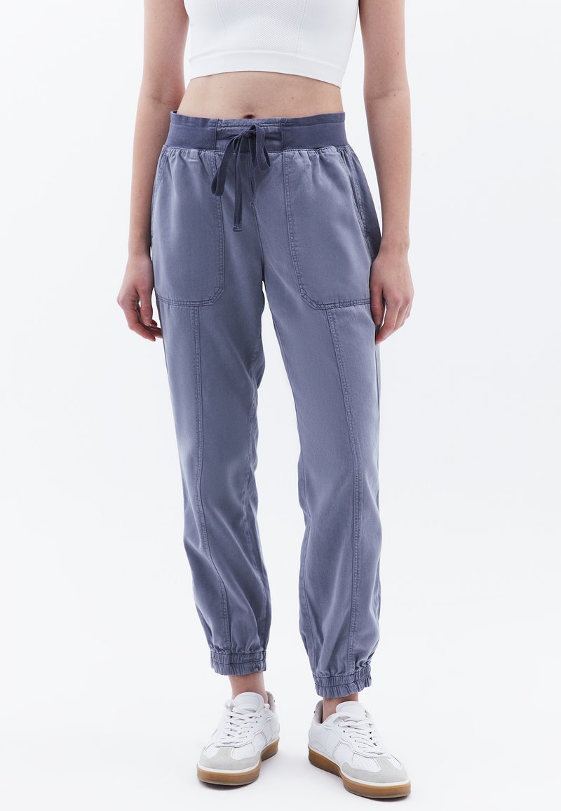 Elastic waist joggerbroeken van een zachte, lichte paarse stof met twee voorzakken, strakke boorden en een verstelbare koord. Rubberzolen van de schoenen zichtbaar.