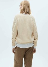 Beige gestreifter Pullover mit entspanntem Schnitt, der über einem weißen Kragenhemd getragen wird, kombiniert mit braunen Hosen. Der Pullover hat überschnittene Schultern und Ballonärmel.