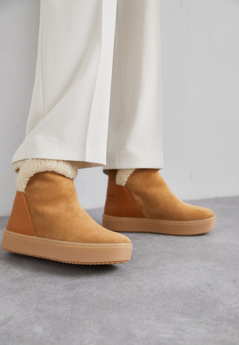 Suede ankle boots in light brown with tan rubber soles, featuring a plush, cream-colored shearling collar. Smooth texture with a sleek design.