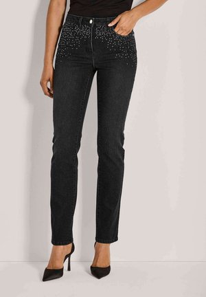 Frau in schwarzen High-Waist-Jeans mit Strassdetails in der Nähe der Taschen, kombiniert mit schwarzen spitzen High Heels.