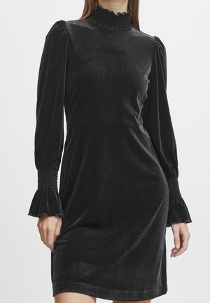 Robe longue en velours noir à manches longues avec col haut à volants et poignets smockés portée par une femme, cadrée à mi-cuisse sur un fond uni.
