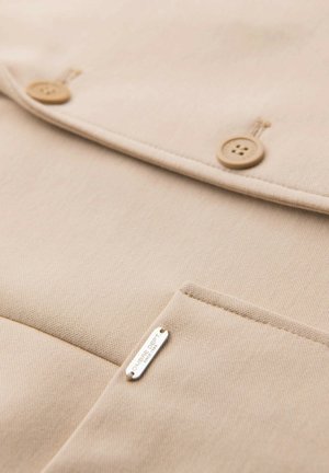 Tissu beige avec deux boutons ronds beiges et une poche comportant une petite étiquette en métal portant la mention "OMBRE DEPT. SINCE 1957."