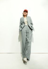 Grijs gestreept pak met een oversized blazer en wijde broekspijpen, gecombineerd met een licht tanktop en een rode pet. Witte sneakers maken de look compleet.