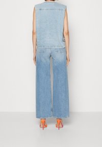 Person som bär en ärmlös ljusblå denimväst och vida jeans med högklackade sandaler i orange och rosa, stående mot en vit vägg.