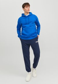 Blue hoodie with a kangaroo pocket, paired with navy joggers featuring white text. The model wears white sneakers. Smooth textile finish.
