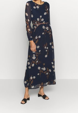 Marineblå chiffonkjole med blomsterprint og lange ærmer, prydet med hvide margueritter og brune grene, indsnøret i taljen med et tyndt bælte. Sorte sko med åbne tæer.