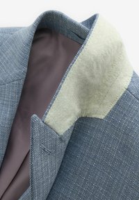 Blauer strukturierter Blazer mit einem leichten, weichen Kragen und grauem Innenfutter. Verfügt über markante Revers und detaillierte Nähte entlang der Kanten.