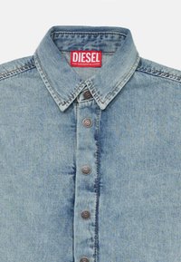 Svetlo modra denimska srajca s klasičnim ovratnikom, petimi gumbi in kontrastnimi šivi. Oznaka nosi napis "DIESEL" v debeli rdeči barvi.