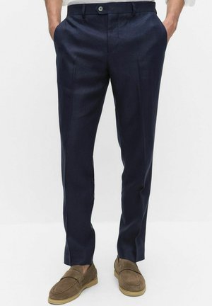 Mężczyzna w dopasowanych granatowych spodniach i brązowych zamszowych mokasynach, z rękami w kieszeniach, na jasnym, jednolitym tle.