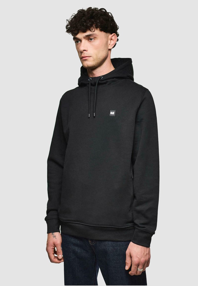 Sweat à capuche noir avec une coupe standard, cordon de serrage ajustable, petite patch logo sur la poitrine, et bords côtes aux poignets et à l'ourlet. Porté avec un jean foncé.