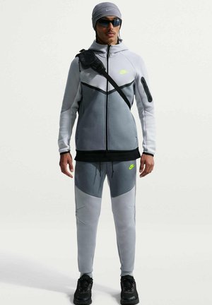 Mężczyzna ubrany w szary i ciemnoszary dres Nike z neonowym logo, czarne trampki, czarna torba na ramię, szara czapka i ciemne okulary przeciwsłoneczne, stojący.