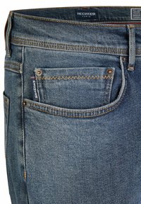 Gros plan sur une poche de jean en denim bleu avec des coutures orange, des rivets en métal, une étiquette de marque et une étiquette "Hechter Paris" sur la taille.