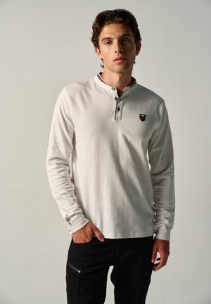 Joven con una camisa polo blanca de manga larga con un pequeño logo en el pecho, de pie con una mano en el bolsillo frente a un fondo liso.
