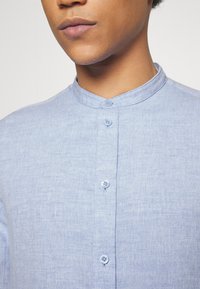 Camisa de lino azul claro con cuello alto, que presenta una tapeta de botones y una textura suave. Ajustada al cuerpo con sutiles detalles de tejido.