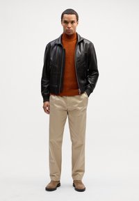 Giacca di pelle nera, maglione a collo alto lavorato arancione, pantaloni beige e stivali in camoscio marroni, in piedi contro uno sfondo semplice.