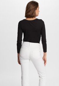 Top nero a maniche lunghe abbinato a jeans bianchi aderenti, con tasche posteriori e vita leggermente alta, su uno sfondo semplice.