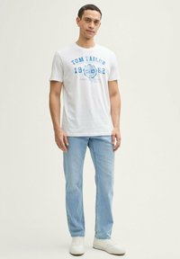 Witte katoenen t-shirt met blauwe grafische print, ronde hals, korte mouwen, gecombineerd met lichtblauwe denim jeans en witte sneakers.