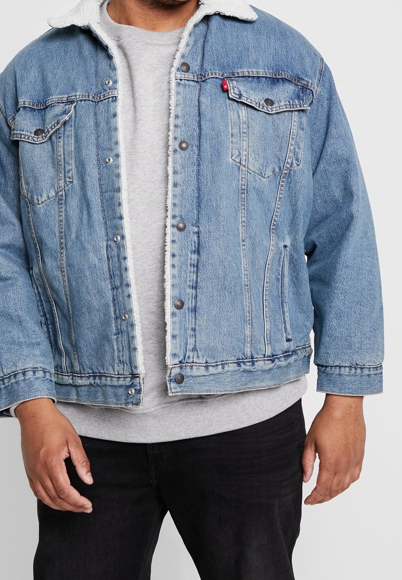 Blekt blå denimjacka med vit fleecekrage, framfickor och tryckknappar. Använd över en grå sweatshirt med rund halsringning.