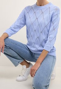 Jasnoniebieski sweter dziewiarski we wzór diamentów z ażurowymi detalami. Posiada okrągły dekolt i rękawy o długości trzy czwarte, zestawiony z niebieskimi dżinsami.