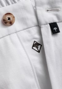 Weißer Stoff mit gewebter Textur, ausgestattet mit einem braunen Knopf und einem gestickten Logo in einem Quadrat. Schwarzes Etikett mit Logo-Akzent inklusive.