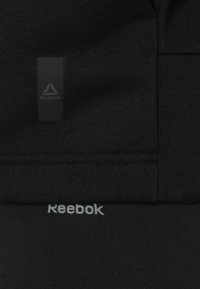 Černá sportovní tkanina Reebok s texturovaným povrchem má matný černý štítek s logem a světle šedý text Reebok poblíž lemu.