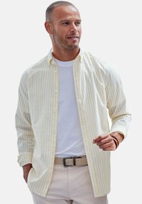 Gestreifte Hemdbluse in Hellgelb und Weiß, mit langen Ärmeln und lockerem Schnitt, kombiniert mit einem weißen T-Shirt und hellbeigen Hosen.