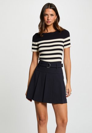 Kvinde med langt mørkt hår iført sort-hvidt stribet top med korte ærmer og sort plisseret nederdel med bælte, stående mod en ensfarvet baggrund.