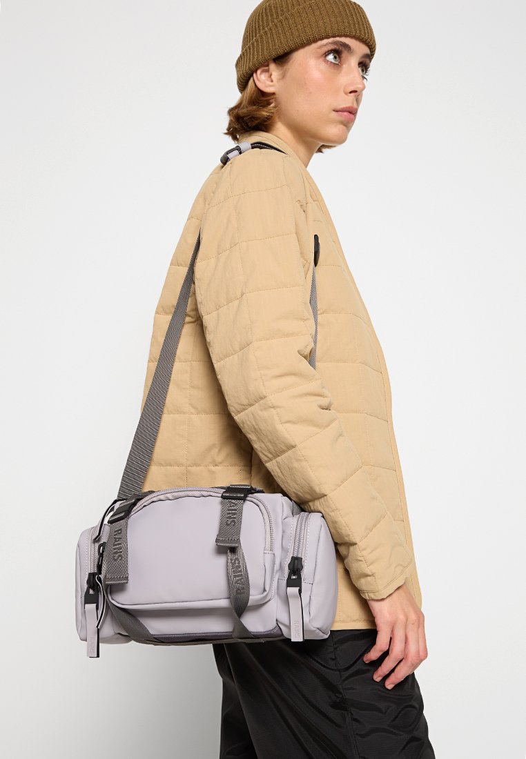 Persoon in een beige gewatteerde jas en een bruine beenhoed die een lichtgrijze crossbody tas met zwarte gespen en banden draagt.