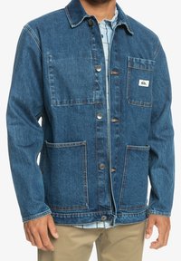 Kurtka jeansowa w ciemnym niebieskim kolorze z zapięciem na guziki, dwoma przednimi kieszeniami oraz naszywką z logo. Charakteryzuje się teksturowanym wykończeniem i kontrastowymi przeszyciami.