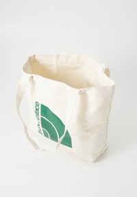 Kreminės spalvos drobės pirkinių maišelis su žalia grafine logotipo. Jis turi du rankenėles ir atidarytą viršų, kad būtų lengva prieiti.