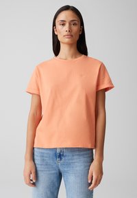 T-shirt di cotone color pesca con collo rotondo, maniche arrotolate e un piccolo simbolo a forma di cuore sul lato sinistro del petto. Indossato con jeans azzurri chiari.