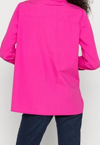 Långärmad skjorta i stark rosa tyg med rak skärning, som har en söm på ryggsidan och böjd fåll. Matchad med mörkblå byxor.