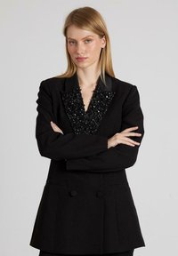 Blazer nero con un colletto a V strutturato e impreziosito, fronte doppiopetto e tessuto liscio. Maniche lunghe e vestibilità sartoriale.