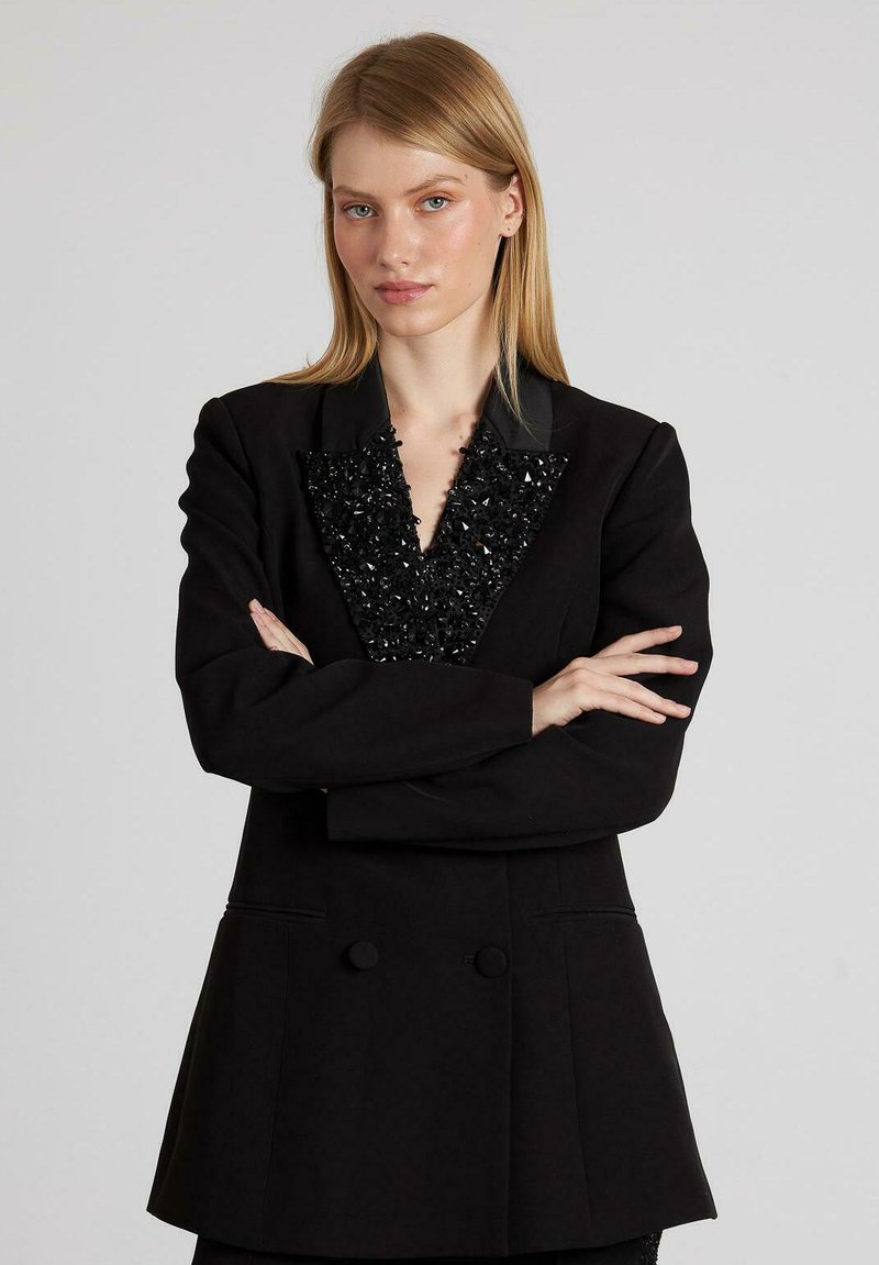 Blazer nero con un colletto a V strutturato e impreziosito, fronte doppiopetto e tessuto liscio. Maniche lunghe e vestibilità sartoriale.