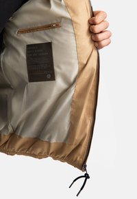 Lekka, beżowa kurtka wewnątrz ma gładką beżową podszewkę, kieszeń na zamek oraz metkę z instrukcją pielęgnacji przymocowaną od wewnętrznej strony.