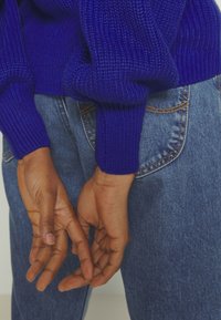 Stickad tröja i livlig blå färg med texturerad tyg, långa ärmar och ribbade ärmslut, kombinerad med ljusblå jeans, med händerna hopknäppta bakom ryggen.