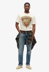Camiseta gráfica de crema con diseño colorido, jeans azules y camisa de cuadros atada. Zapatillas beige con suelas texturizadas. Atuendo casual.