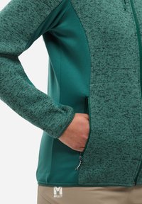 Personne portant une veste zippée texturée verte avec une main dans la poche zippée latérale, mettant en évidence le détail de la manche et la texture du tissu.