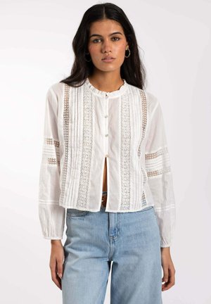 Blusa blanca de botones con un cuello con volantes, inserciones de encaje y detalles plisados. Mangas largas y un corte cropped. Combinada con jeans de color azul claro.