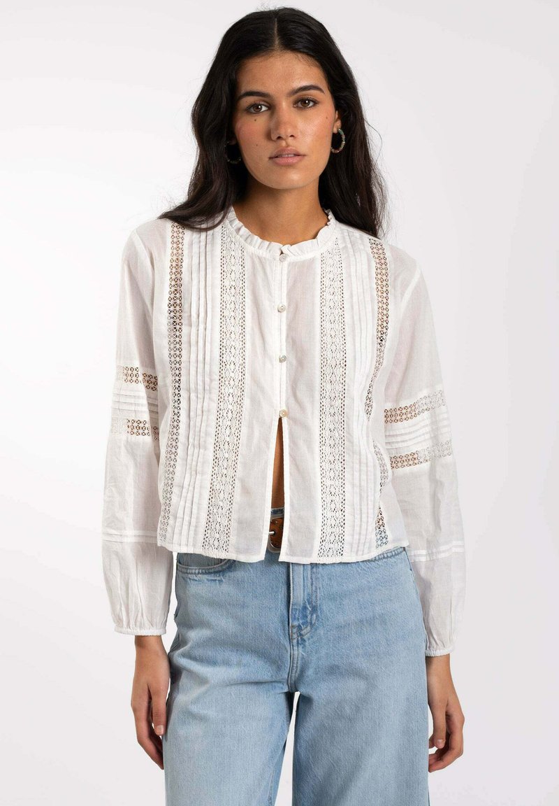 Blusa blanca de botones con un cuello con volantes, inserciones de encaje y detalles plisados. Mangas largas y un corte cropped. Combinada con jeans de color azul claro.