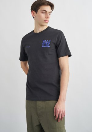 Ung mann iført svart Nike T-skjorte med blå gotisk logo på brystet, kombinert med olivengrønne bukser, står mot en ensfarget grå bakgrunn.