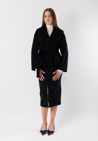 Giacca avvolgente nera in lana con ampio colletto, vita cintata e maniche lunghe, abbinata a una gonna nera al ginocchio con uno spacco frontale. Tacchi neri.