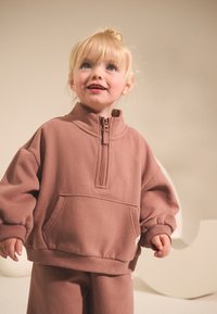 Maglione in pile marrone con collo alto, mezza zip, taglio oversize, polsini a costine e tasca frontale. Abbinato a pantaloni marroni coordinati.