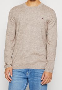 Beige stickad tröja med rund hals och långa ärmar; har en liten logotyp på vänster bröst. Buren över blå jeans.
