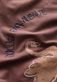 Tkanina z haftowanym niebieskim napisem "WELCOME FAR NUENTE" oraz nadrukiem brązowego łabędzia na wrzosowym tle.